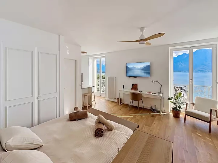 Pièce principale lumineuse avec lit, bureau et portes-fenêtres ouvrant sur un balcon avec vue sur le lac
