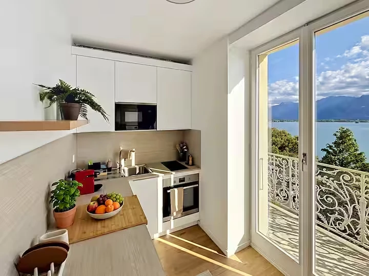 Cuisine blanche équipée ouverte sur une porte-fenêtre et un balcon donnant sur le lac et les montagnes